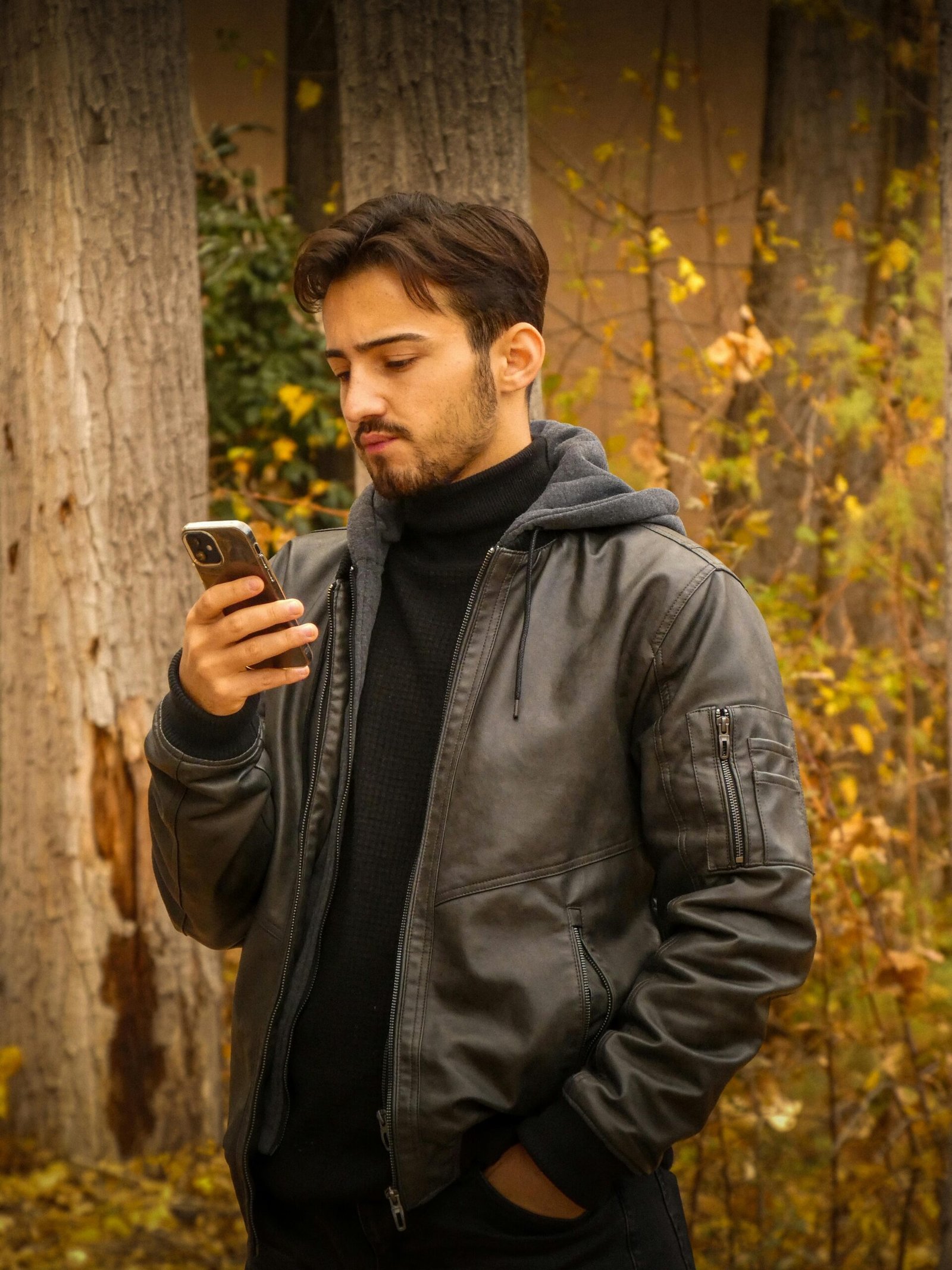 A man in a leather jacket checks his smartphone in a serene autumn forest.